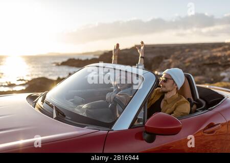 Joyful couple enjoying vacations, driving together convertible car on the rocky ocean coast on a sunset. Happy vacation, love and travel concept Stock Photo