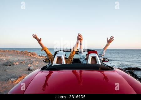 Couple driving convertible car, traveling near the ocean on a sunset, view from the backside. Happy vacations and traveling by car concept Stock Photo