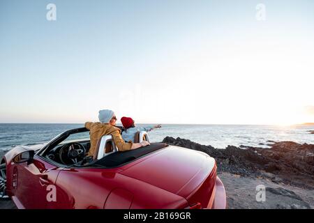 Couple enjoying beautiful view on the ocean, traveling by convertible car near the ocean on a sunset, view from the backside. Happy vacations and traveling by car concept Stock Photo