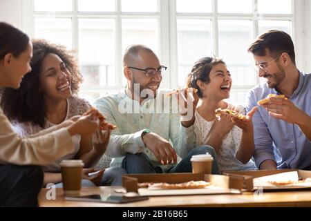 Five laughing multi-ethnic friends eating pizza indoors Stock Photo