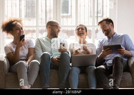Cheerful multiracial friends using diverse gadgets sitting together on couch Stock Photo