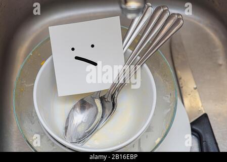 Some dirty dishes, spoons and knife in iron sink and paper with sad face, concept picture Stock Photo