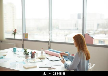 Wide angle portrait of red haired female designer holding color swatches while sitting at workplace by window, copy space Stock Photo