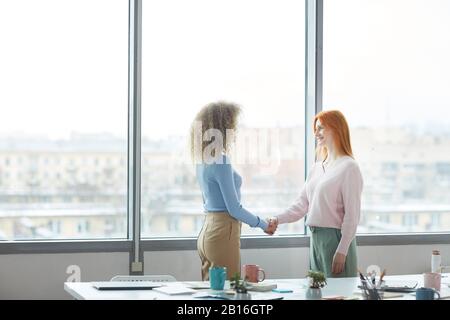 Side view portrait of two successful young businesswomen shaking hands after job interview or deal while standing by window in modern office, copy space Stock Photo
