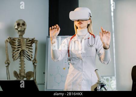 Young woman doctor or scientist wearing VR headset glasses device working with virtual reality touch screen. Modern lab with human skeleton on the bac Stock Photo