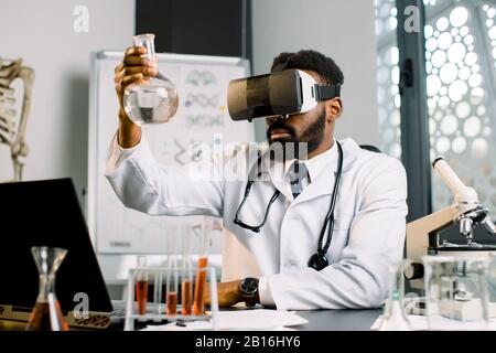 Young African man lab worker wearing VR goggles at work. Science technician in virtual reality glasses examines the drug in laboratory, holding glass Stock Photo