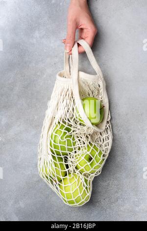 Hand holding mesh bag with green organic apples over grey concrete background. Concept of zero waste, reusable shopping bag, sustainable lifestyle Stock Photo
