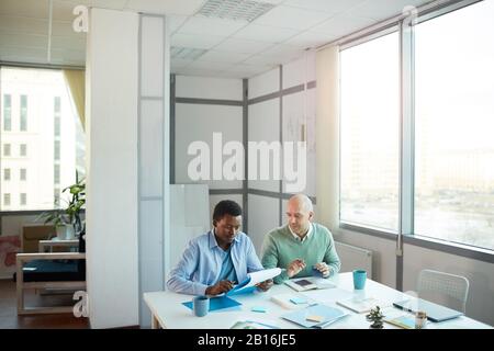 Wide angle view of African-American young man consulting with mature manager while sitting at table in office, copy space Stock Photo