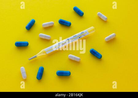 Mercury thermometer, white and blue capsules isolated on bright yellow background. Epidemic, painkillers, healthcare and treatment concept. Flat lay. Stock Photo
