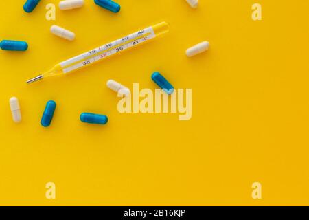 Mercury thermometer, white and blue capsules isolated on bright yellow background. Epidemic, painkillers, healthcare and treatment concept. Flat lay. Stock Photo