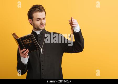thoughtful catholic priest holding bible and looking at wooden rosary beads isolated on yellow Stock Photo