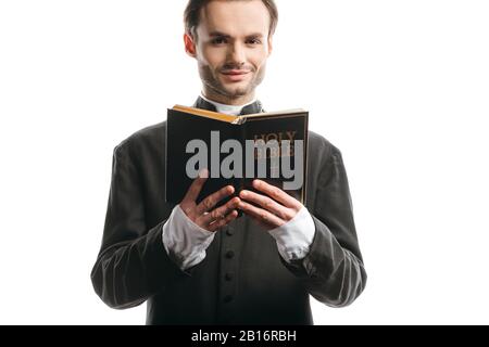 young, smiling catholic priest holding holy bible while looking at camera isolated on white Stock Photo