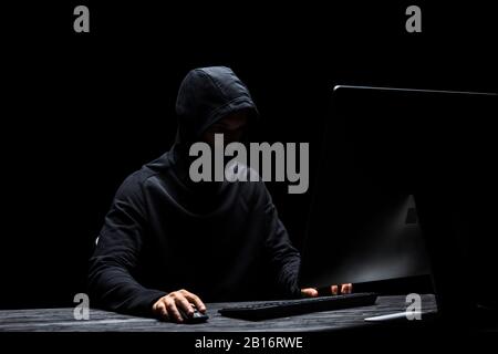 hooded anonymous hacker in mask using computer isolated on black Stock Photo