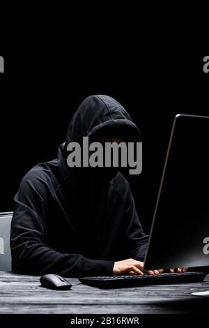 hooded anonymous hacker in mask typing on computer keyboard isolated on black Stock Photo
