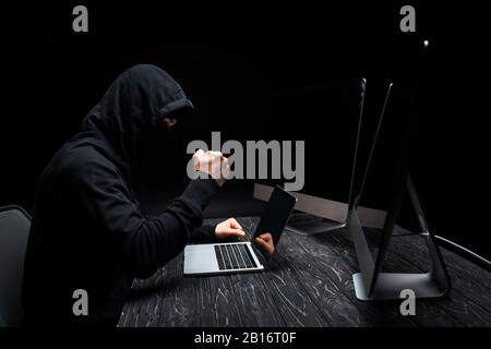 angry hacker with clenched fist near laptop and computer monitors with blank screen isolated on black Stock Photo