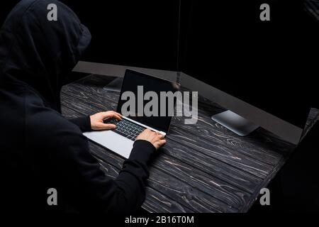 hooded hacker using laptop near computer monitors with blank screen isolated on black Stock Photo