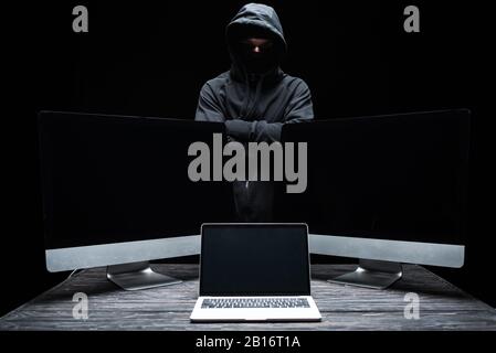 hacker in mask standing with crossed arms near computer monitors and laptop with blank screen  isolated on black Stock Photo