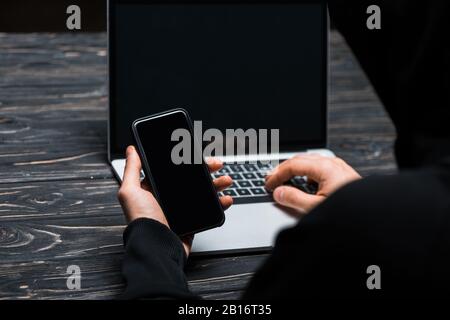 cropped view of hacker using smartphone near laptop with blank screen isolated on black Stock Photo