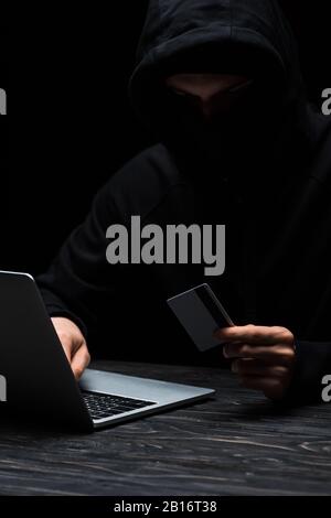 hacker in hood and mask using laptop while holding credit card isolated on black Stock Photo