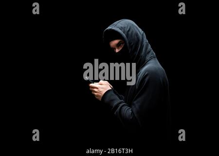 hacker in hood and mask holding smartphone and looking at camera isolated on black Stock Photo