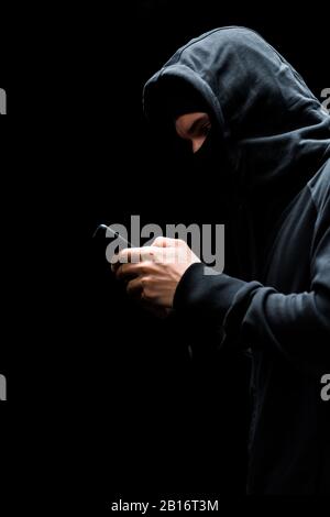 anonymous hacker in hood and mask using smartphone isolated on black Stock Photo