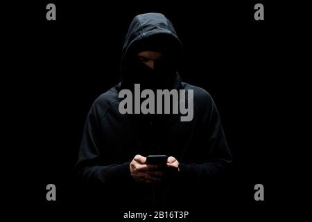 hacker in hood and mask using smartphone isolated on black Stock Photo