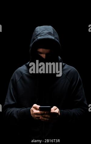 hacker in hood using smartphone isolated on black Stock Photo