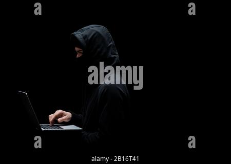 anonymous hacker in hood and mask using laptop isolated on black Stock Photo