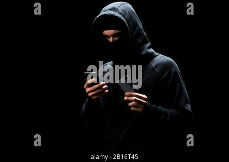 hacker in mask using smartphone and holding credit card isolated on black Stock Photo