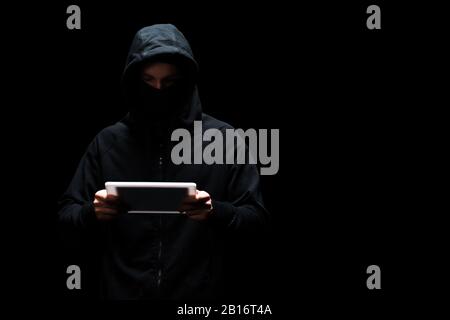 anonymous hacker in hood and mask using digital tablet isolated on black Stock Photo