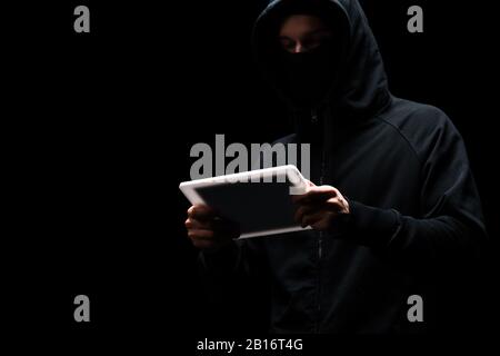 anonymous hacker in hood using digital tablet isolated on black Stock Photo
