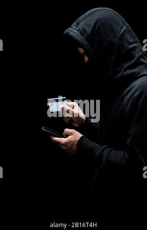 hacker in hood and mask using smartphone and holding credit card isolated on black Stock Photo