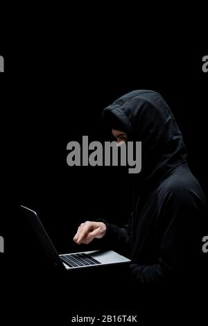 hacker in hood and mask pointing with finger at laptop isolated on black Stock Photo