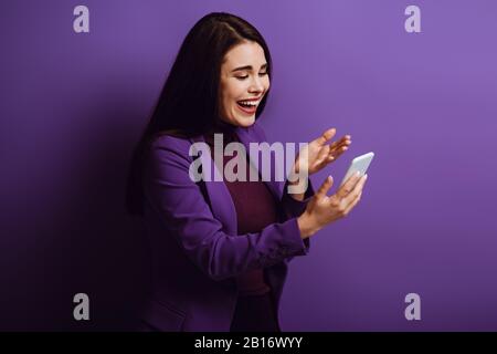 excited girl standing with open arm and laughing while having video chat on purple background Stock Photo