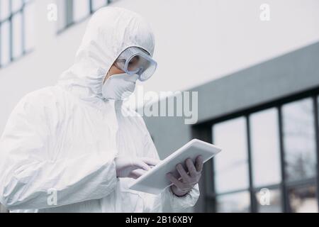 asian epidemiologist in hazmat suit and respirator mask using digital tablet while standing on street Stock Photo