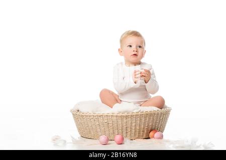 Child holding Easter egg, sitting in basket with colorful decoration around on white background Stock Photo