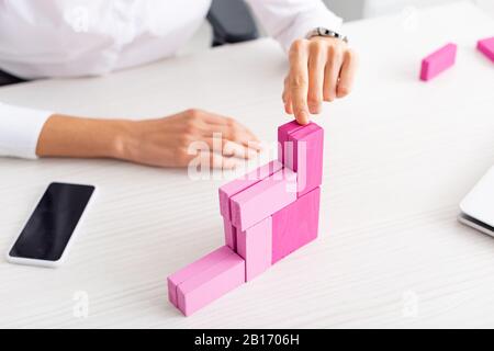 Cropped view of businesswoman stacking marketing pyramid from pink building blocks near gadgets on table Stock Photo