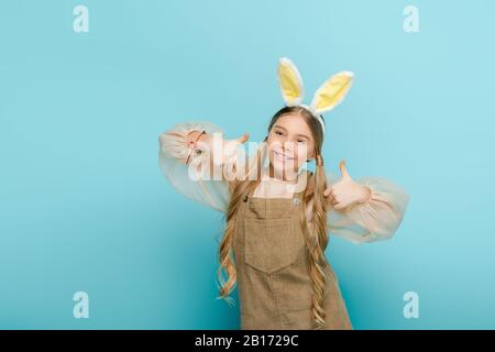 cheerful kid with bunny ears showing thumbs up isolated on blue Stock Photo