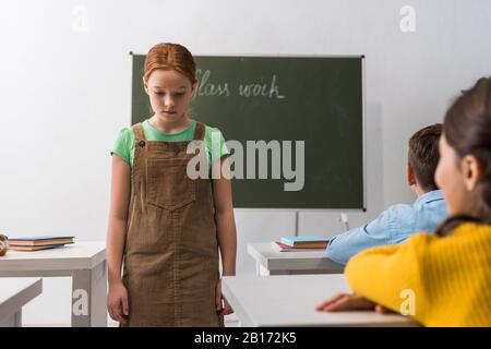 selective focus of upset schoolgirl walking near classmates sitting at desks Stock Photo