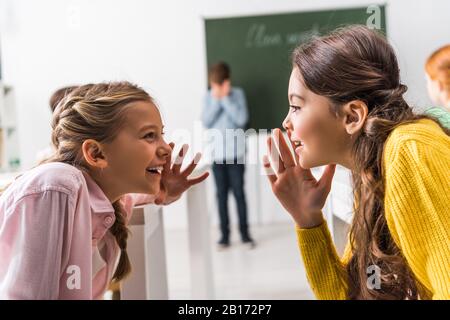 selective focus of schoolgirls gossiping near upset classmate, bullying concept