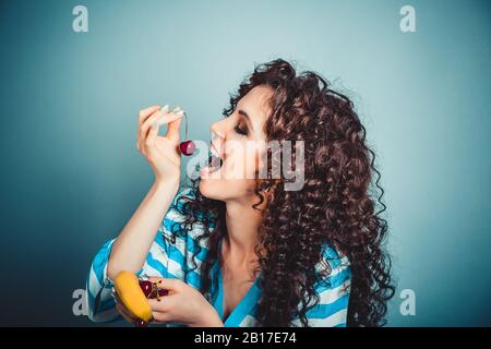 Healthy eating concept. Attractive young beautiful curly woman holding enjoying cherry in open mouth with eyes closed holding banana in other hand hap Stock Photo