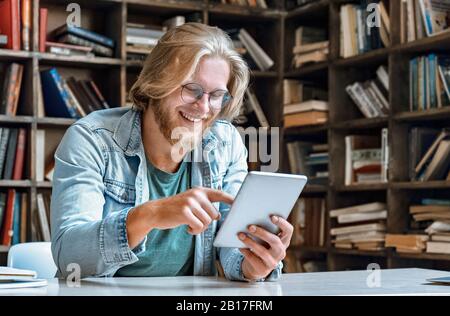 Young bearded man glasses library desk hold use modern tablet laugh at screen Stock Photo