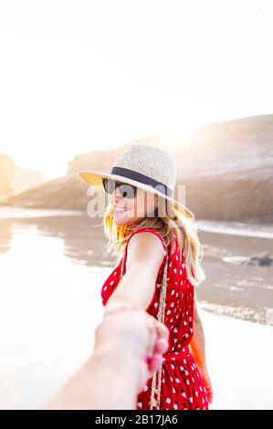 Blond woman wearing red dress and hat and holding hand on the beach, Natural Arch at Playa de Las Catedrales, Spain Stock Photo