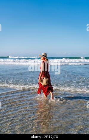 Blond woman wearing red dress and hat and walking at the beach, Playa de Las Catedrales, Spain Stock Photo