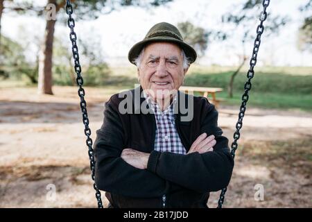 Old man sitting on swing in park, with arms crossed Stock Photo