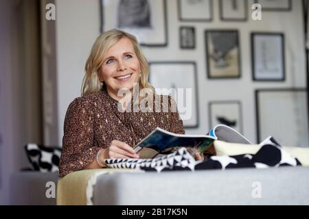 Smiling blond woman relaxing at home sitting on couch Stock Photo