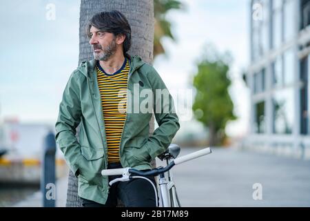 Portrait of serious mature man with  bike leaning against palm tree trunk looking at distance, Alicante, Spain Stock Photo