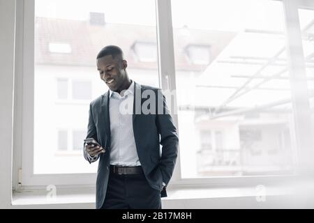 Portrait of smiling young businessman standing in front of window looking at cell phone Stock Photo