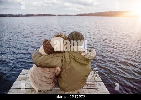 Back view of young couple in love sitting on a jetty looking at lake in winter Stock Photo