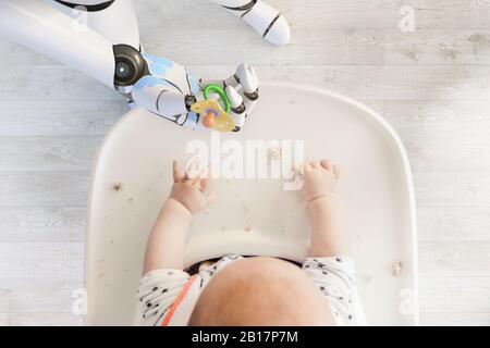 Robot hand giving pacifier to baby boy sitting in high chair playing with bread crumps, top view Stock Photo